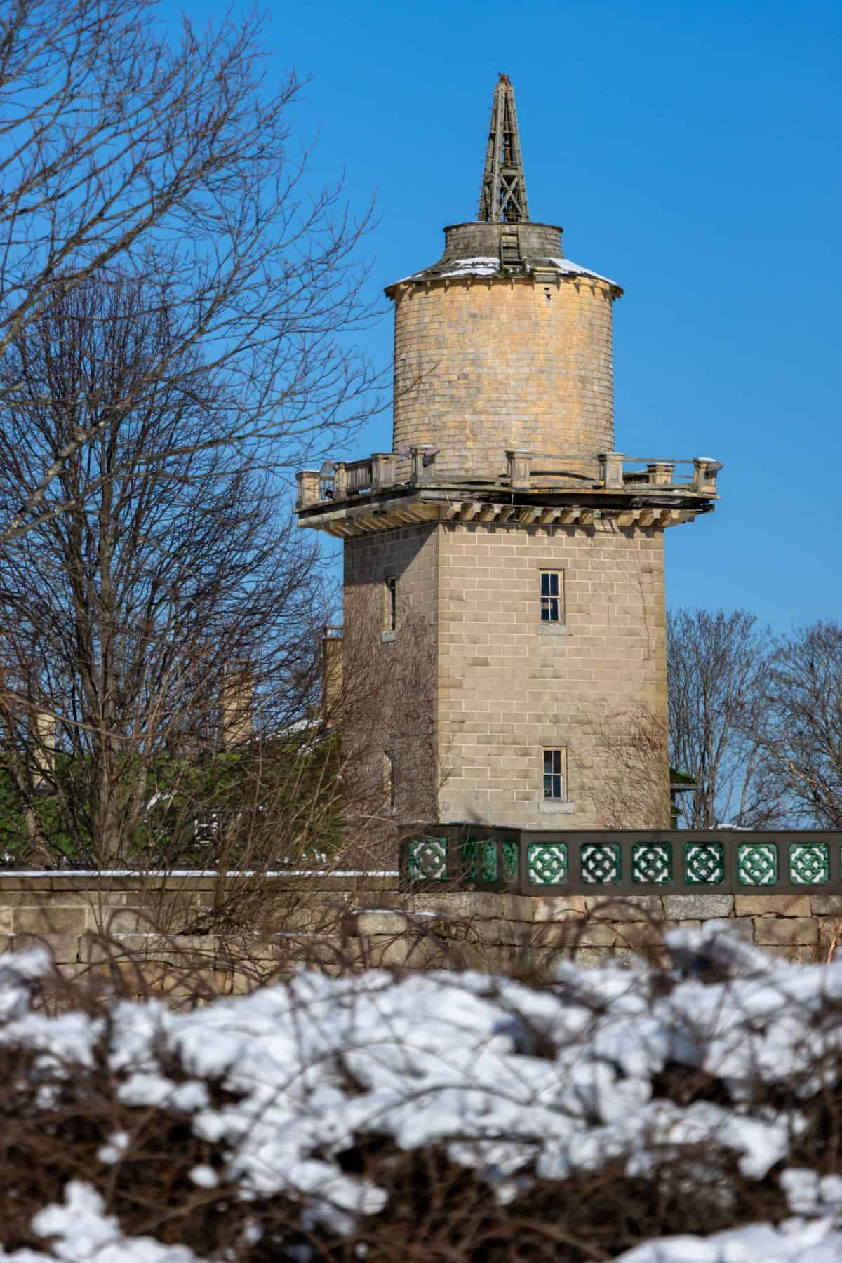A stone tower with a round top section and a pointed structure, surrounded by bare trees and snow-covered bushes, under a clear blue sky.
