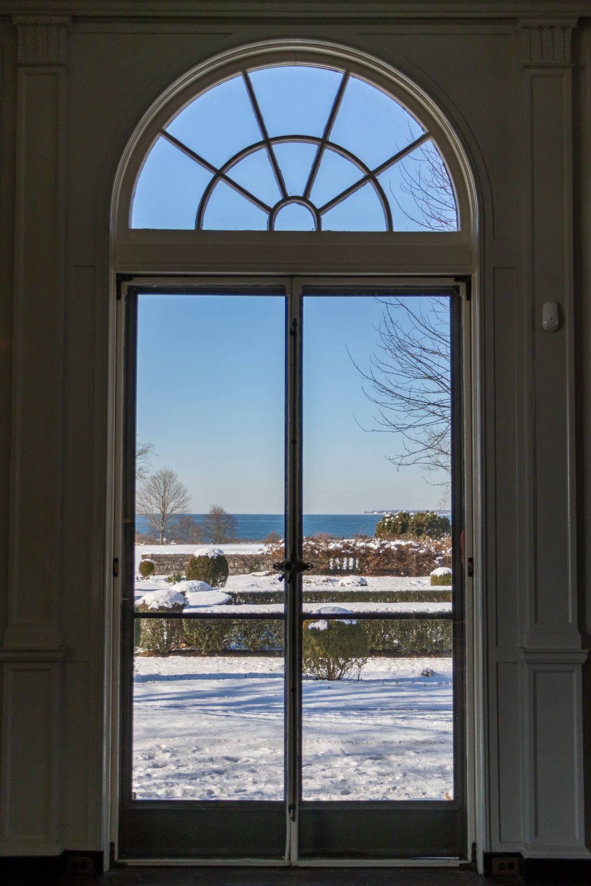 A large glass door with an arched window above opens to a snowy landscape with trees, shrubs, and a distant view of water under a clear blue sky.