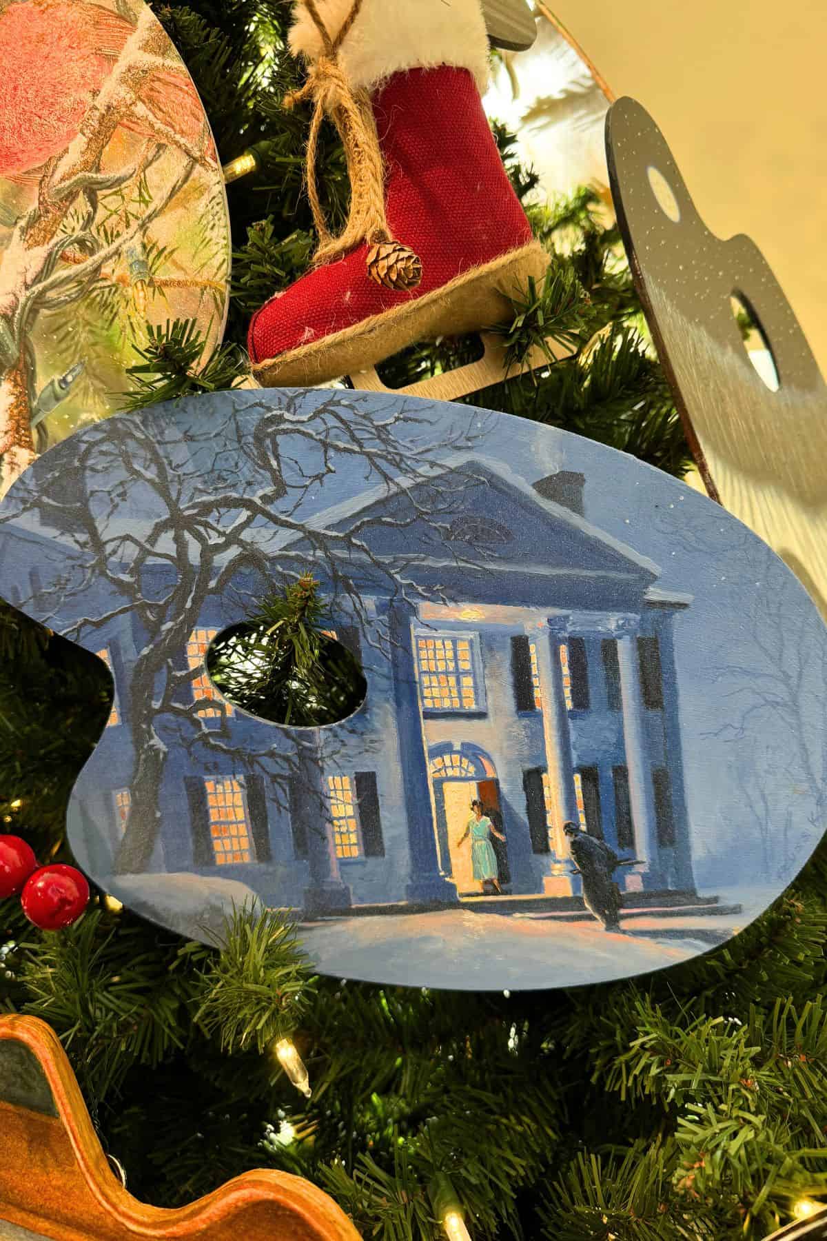 A painter’s palette ornament painted with a wintry scene of a lit-up mansion hangs on a Christmas tree near a small red boot decoration.