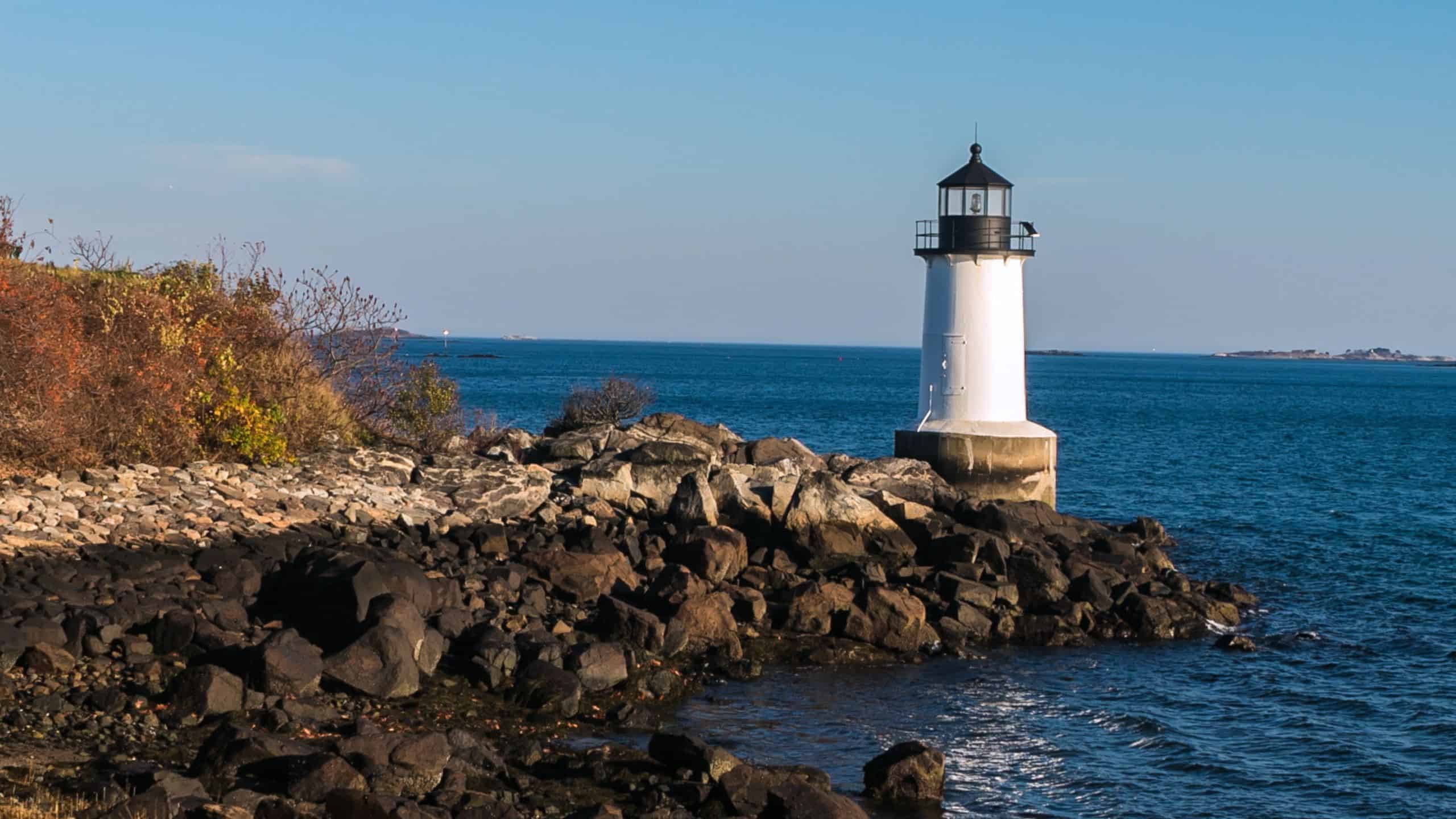 A white lighthouse stands on a rocky shoreline, overlooking a calm blue sea under a clear sky.