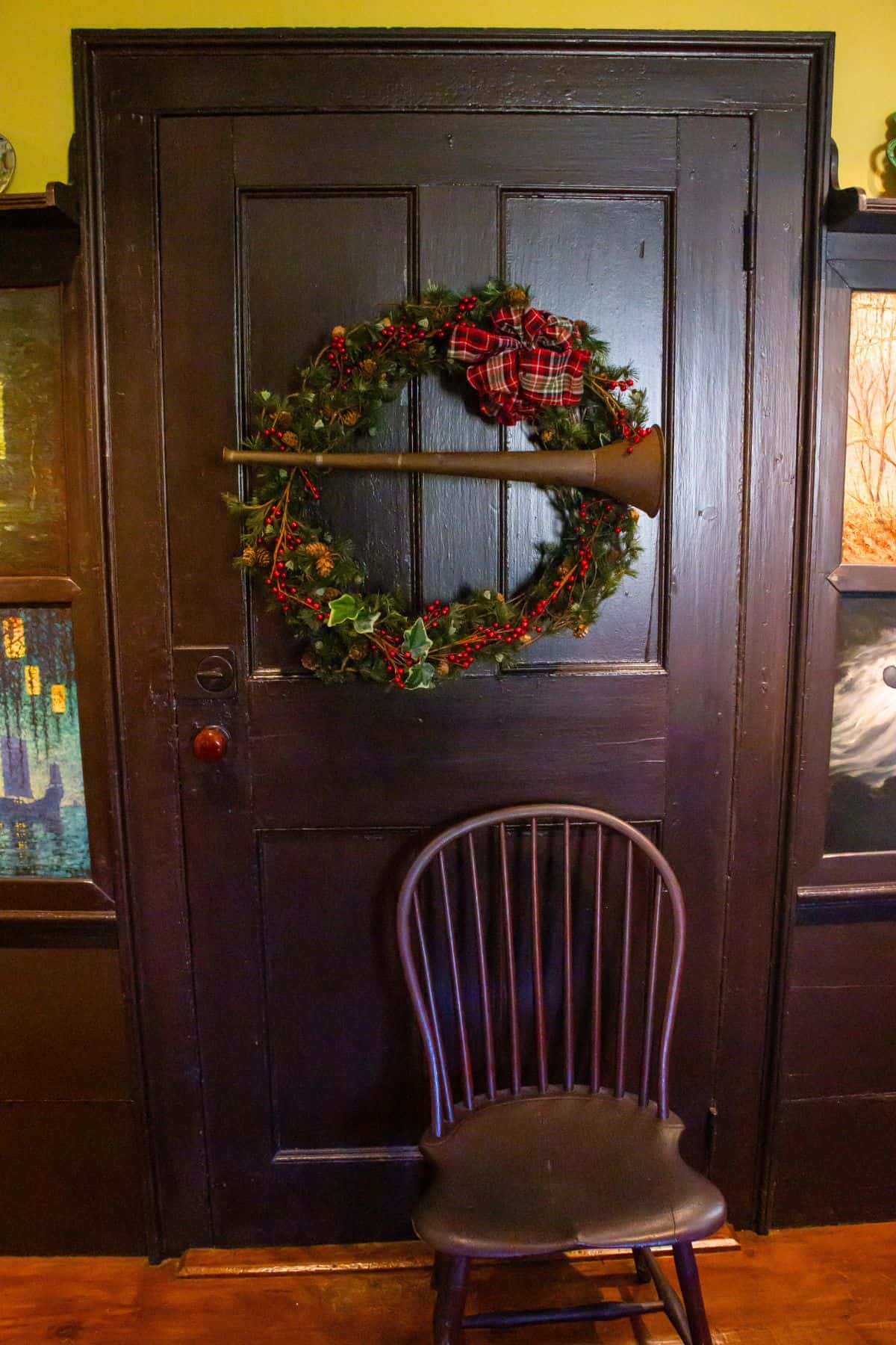 A dark wooden door decorated with a holiday wreath and a horn; a wooden chair is placed in front of the door.