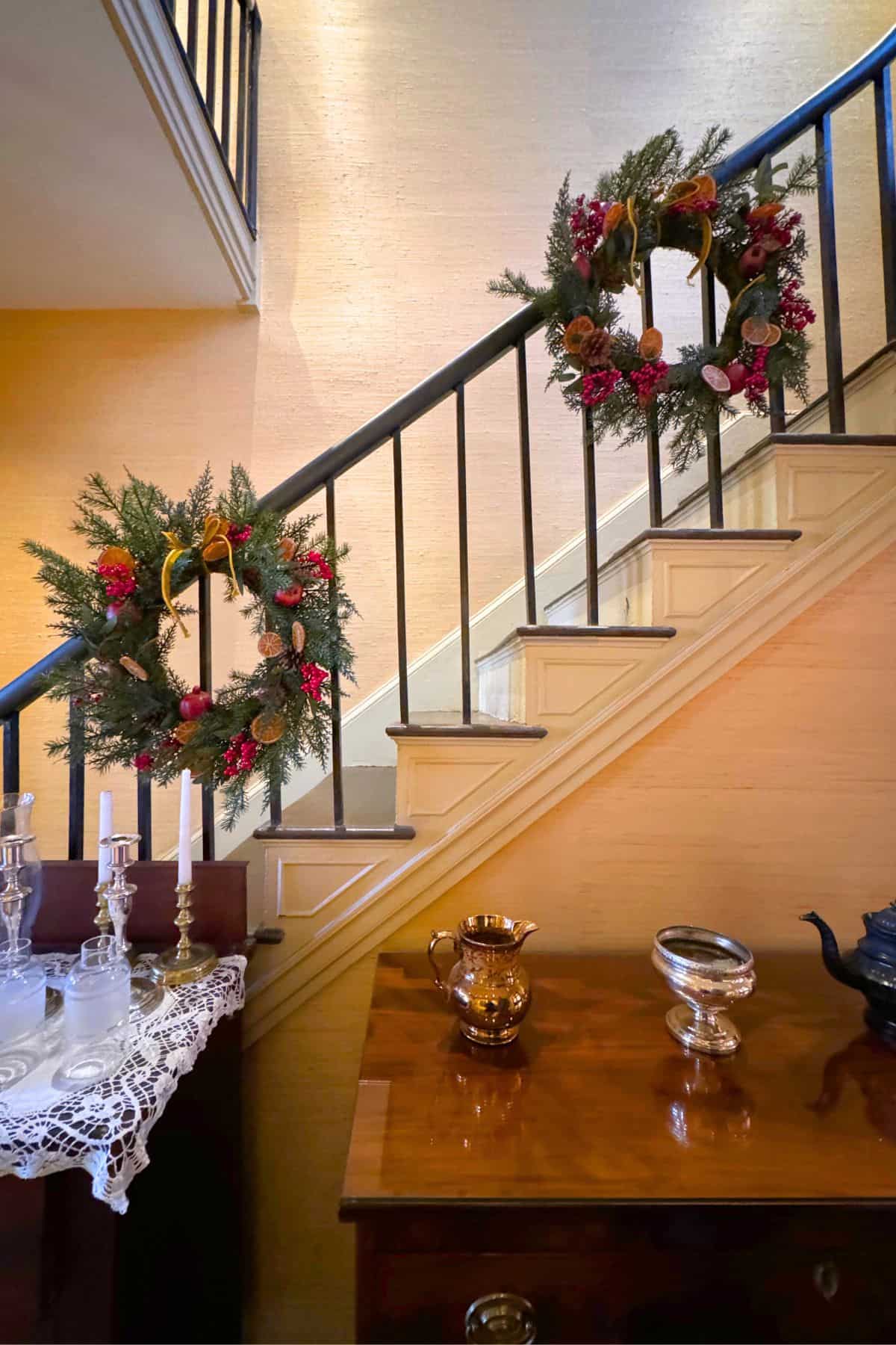 A staircase decorated with two holiday wreaths; in the foreground, a wooden table holds candlesticks, a lace runner, a teapot, and a copper pitcher.
