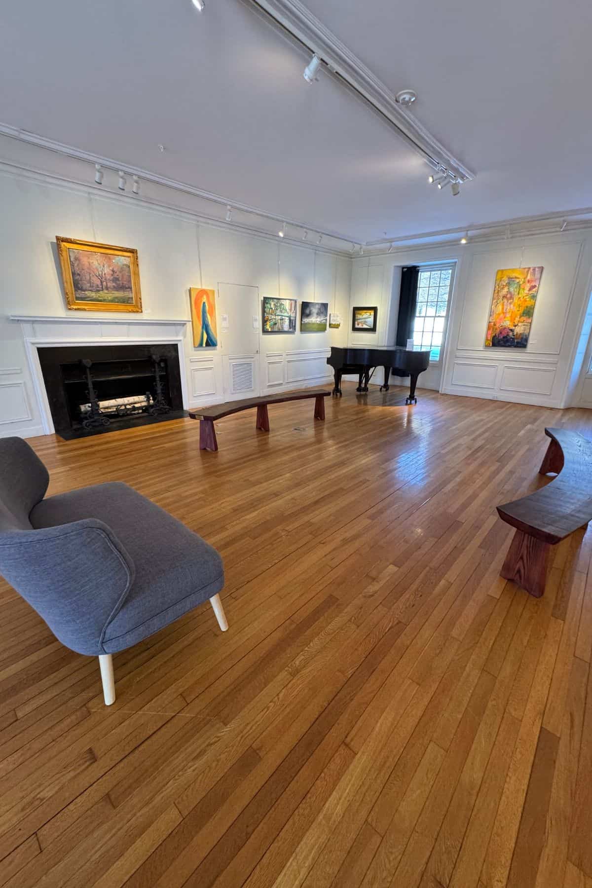Art gallery room with wooden floor, benches, a gray couch, a piano, and several colorful paintings displayed on white walls.