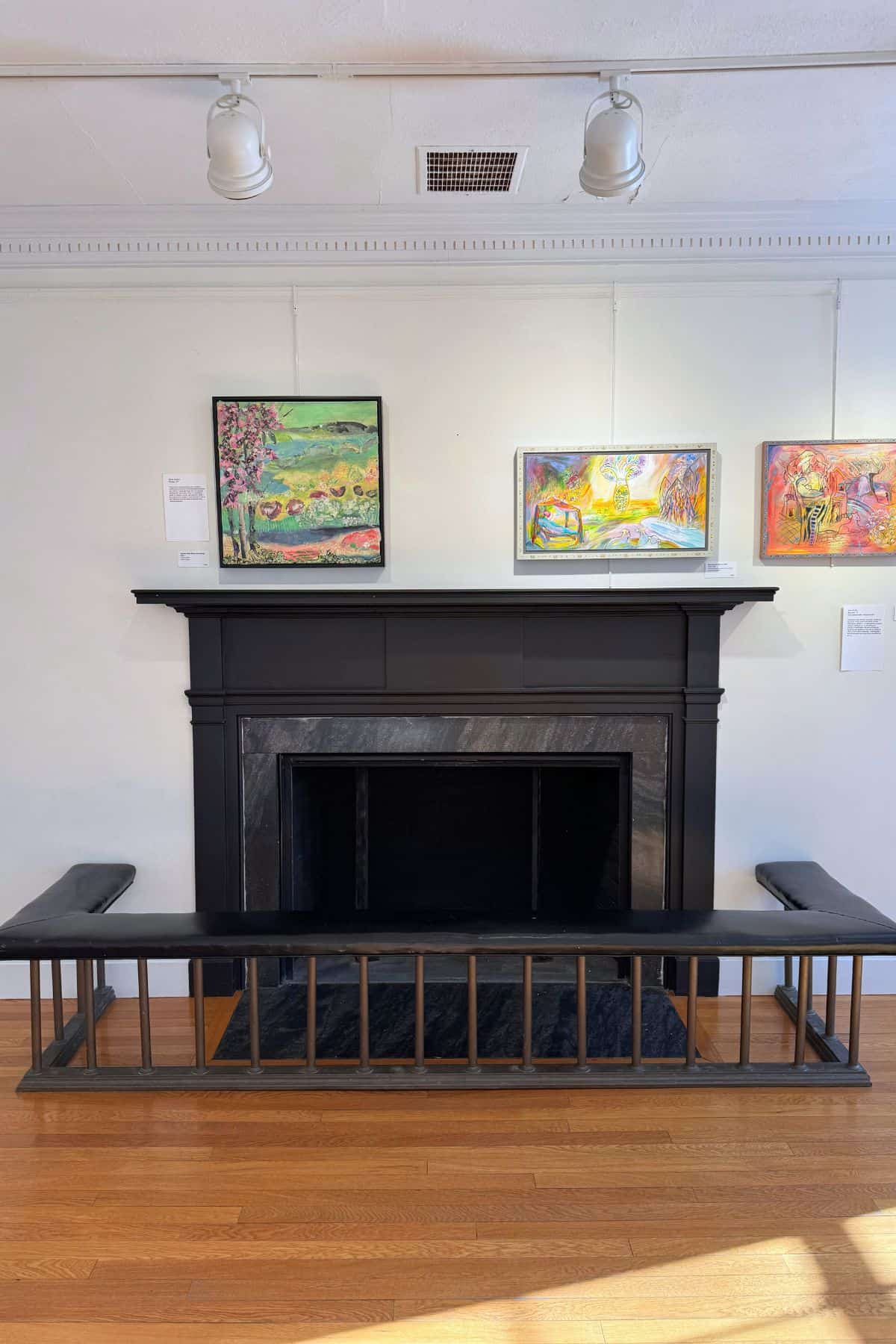 A black fireplace with a surrounding bench sits against a white wall, above which three colorful paintings are displayed in a well-lit room with wood flooring.