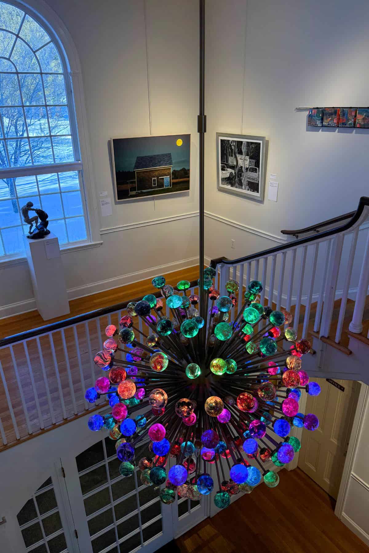 Colorful chandelier with round glass bulbs hangs in an art gallery with paintings on the walls, a large arched window, and wooden floors.