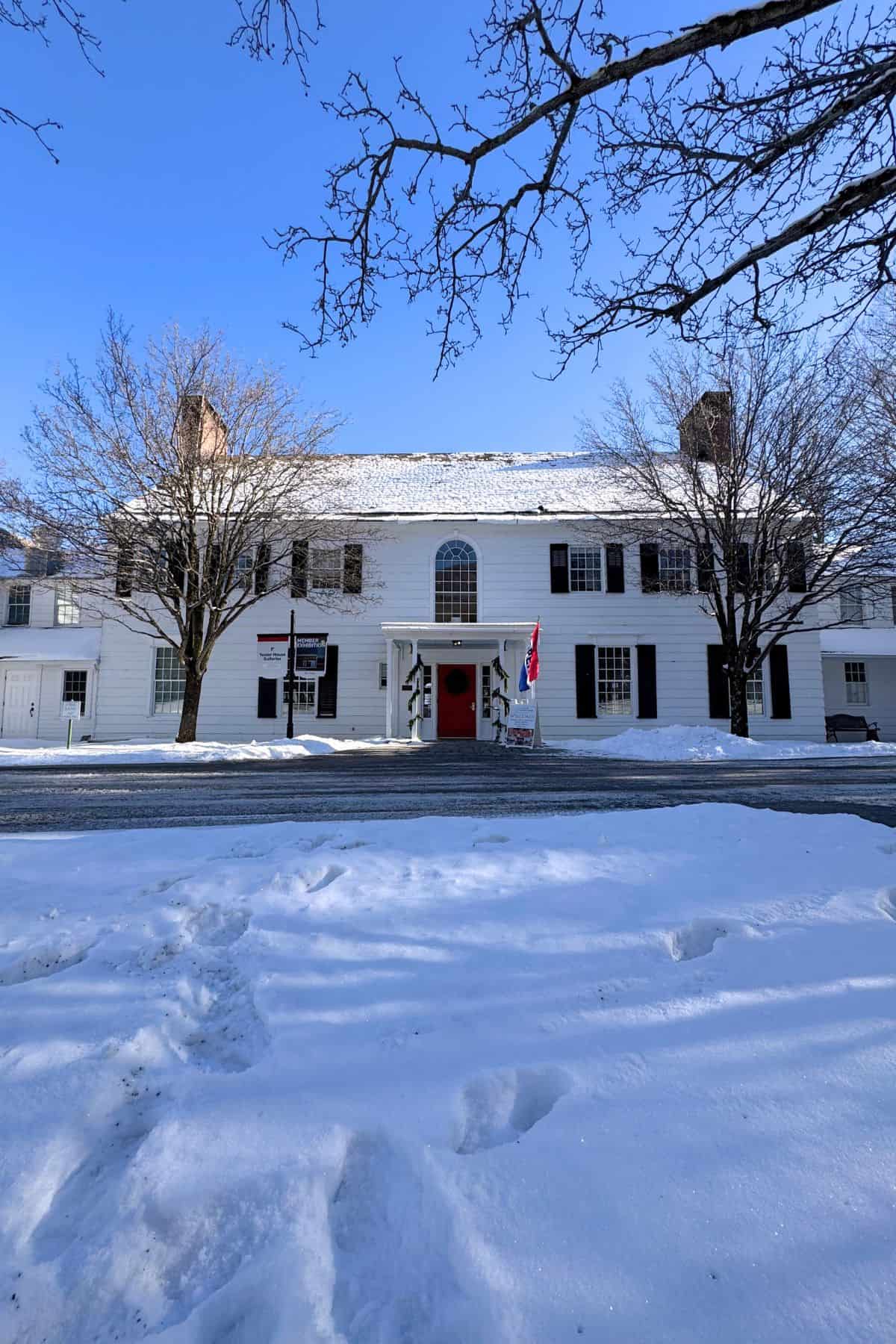 A large white colonial-style building with black shutters and a red door, surrounded by snow and leafless trees under a clear blue sky.