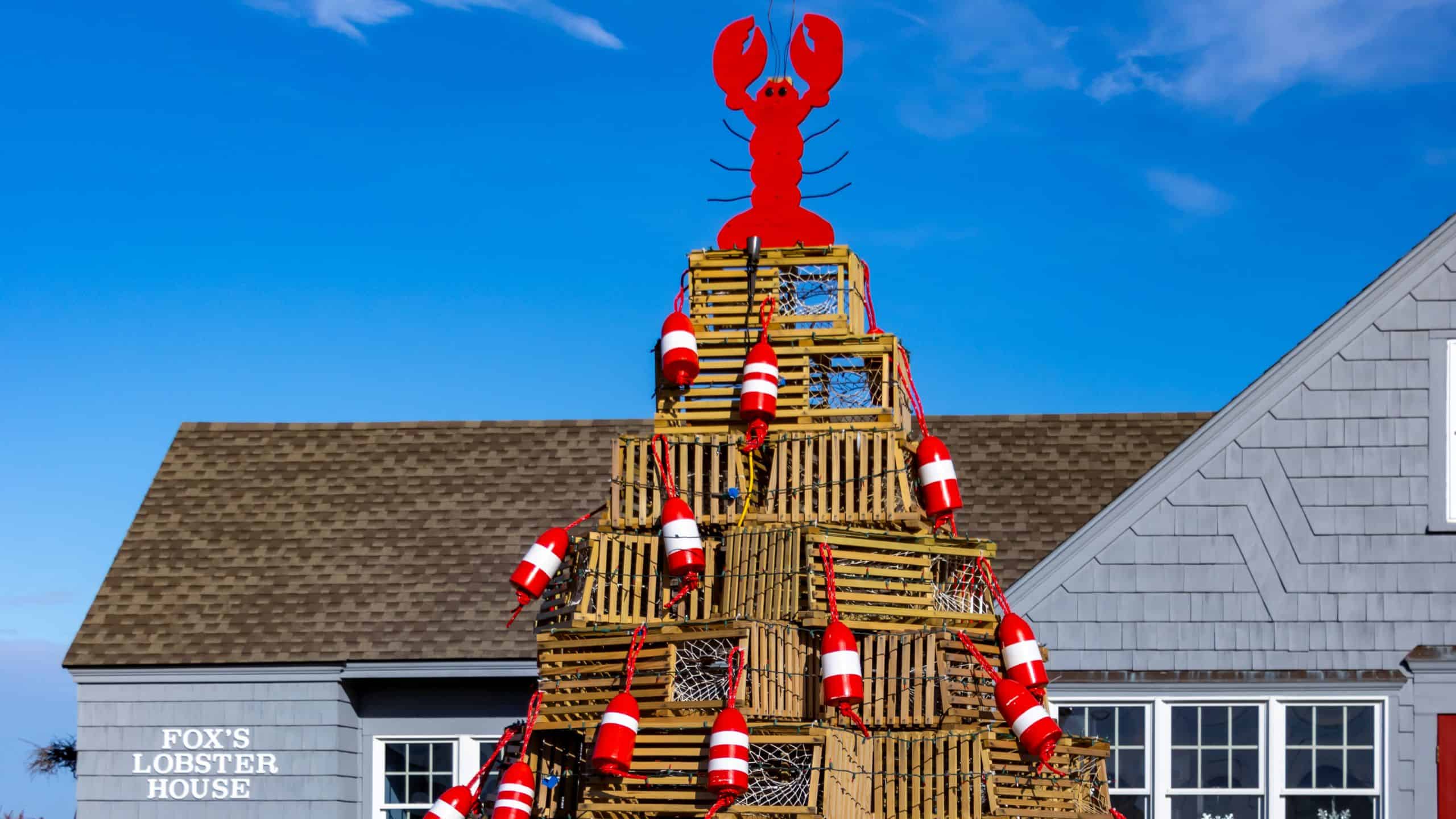 A pyramid-shaped stack of lobster traps decorated with red and white buoys, topped with a red lobster cutout, stands in front of a gray building labeled "FOX'S LOBSTER HOUSE.
