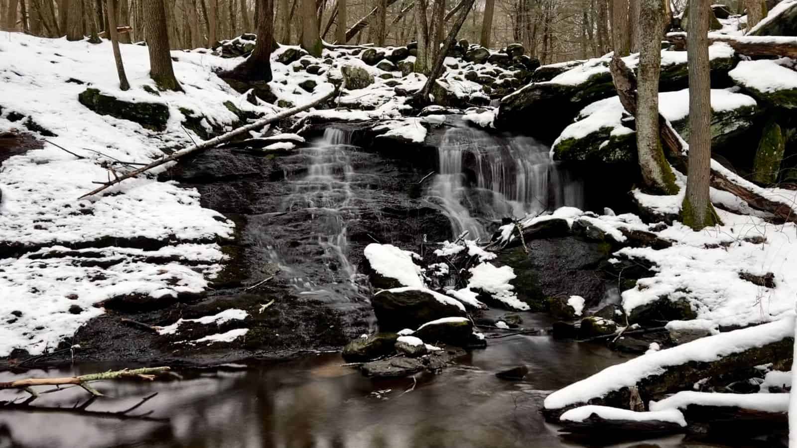A small waterfall flows over dark rocks in a forest with snow on the ground and trees, reflecting in a calm pool below.