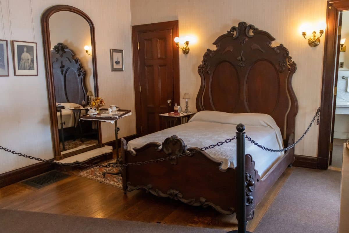 Victorian-style bedroom with a carved wooden bed, mirror, small table, artwork on the walls, and a rope barrier separating the room from visitors.