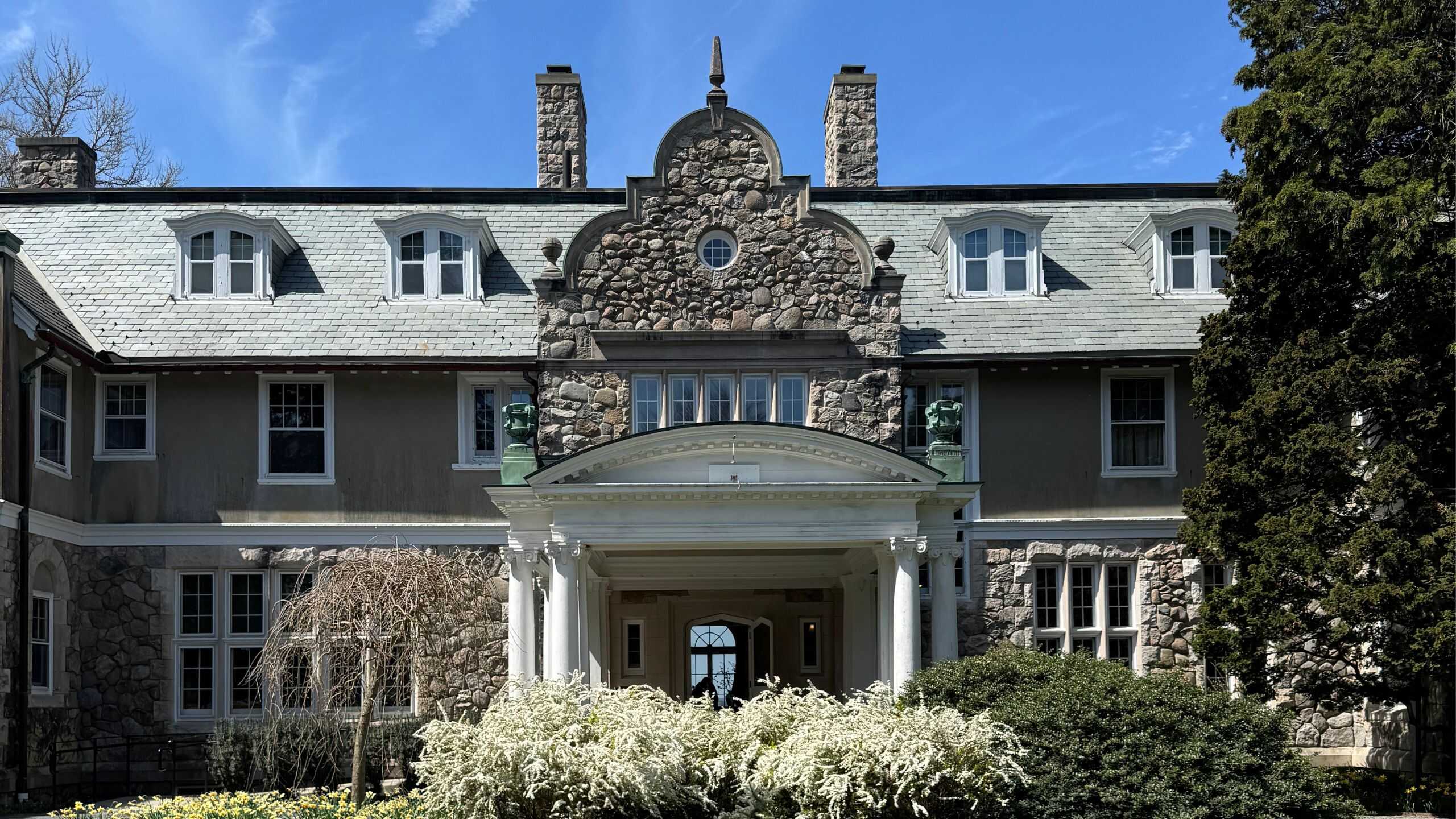 Large historic stone mansion with a central arched pediment, multiple chimneys, gray roof, and a columned entrance surrounded by shrubs and trees under a blue sky.
