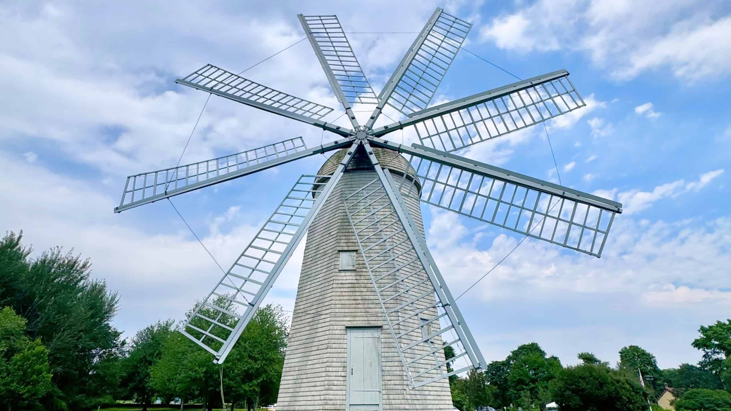 A traditional white windmill with large blades stands on a grassy field, surrounded by trees under a partly cloudy sky.