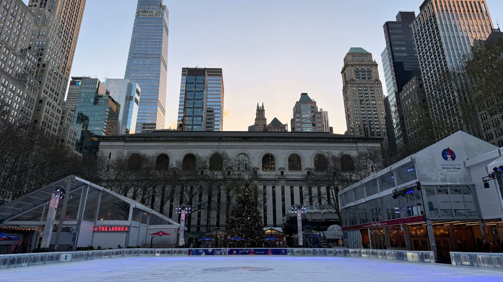 An outdoor ice skating rink with a Christmas tree in the center, surrounded by tall buildings in a city at dusk.