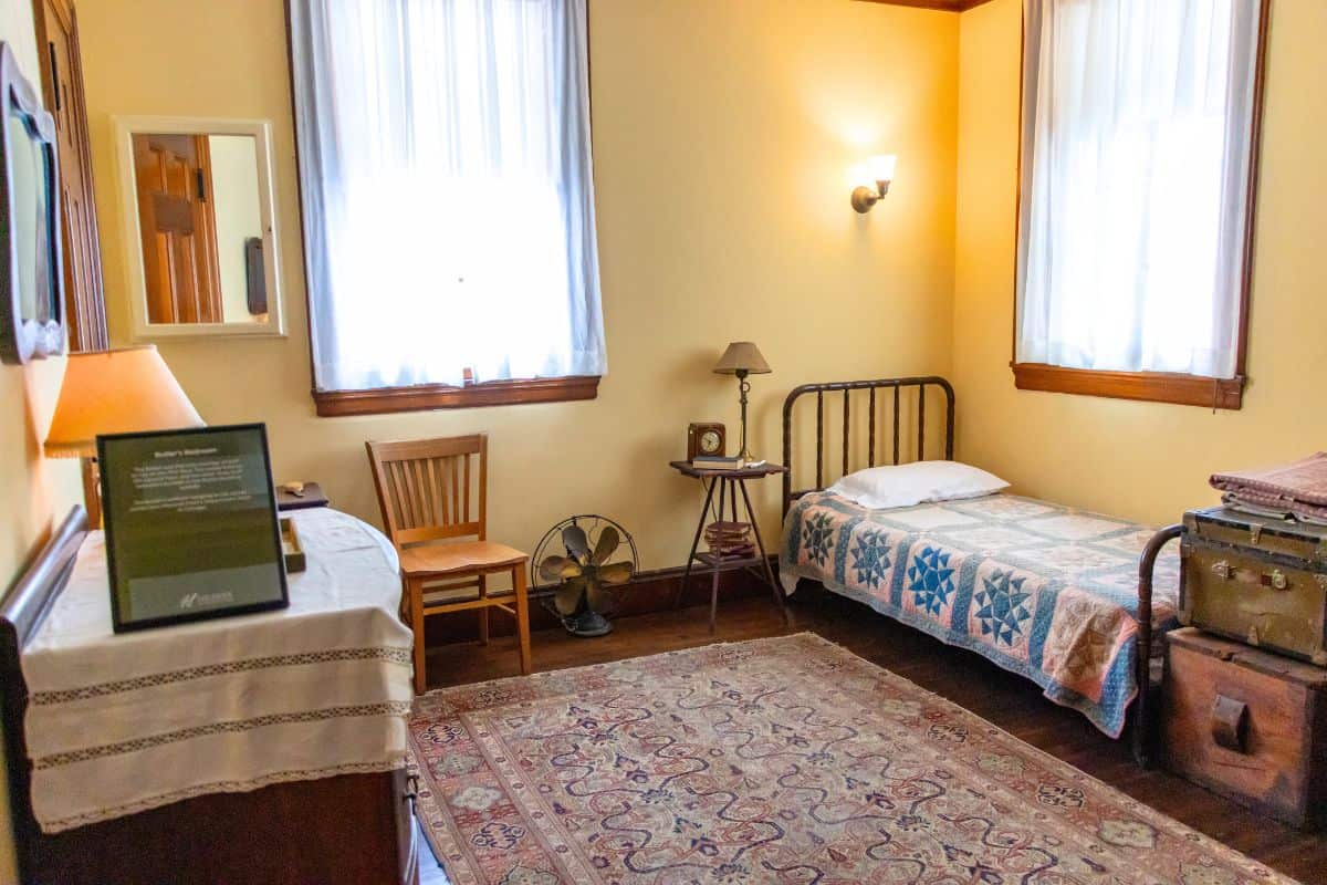 A small, tidy bedroom with a single bed, wooden chair, table, vintage fan, stacked suitcases, and two curtained windows letting in natural light.