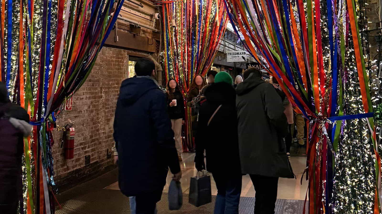 People walk through a corridor decorated with colorful ribbons and lit Christmas trees inside a brick-walled building.