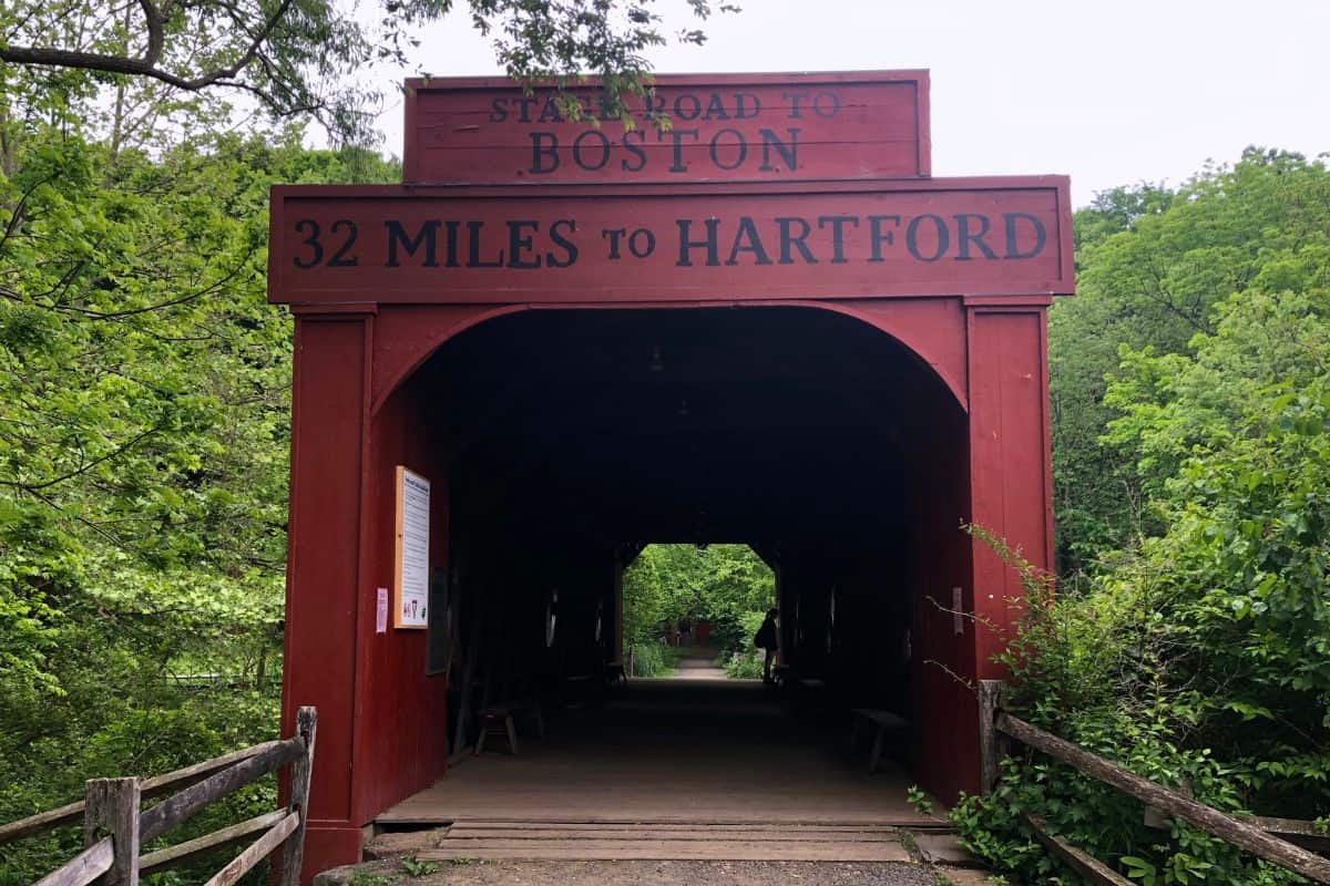 A red covered bridge displays signs reading "State Road to Boston" and "32 Miles to Hartford," surrounded by green trees and a wooden fence.