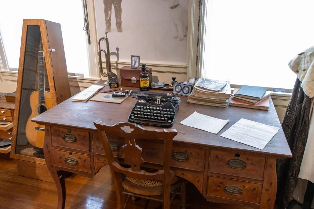 A wooden desk with a typewriter, papers, books, and various objects sits in front of two windows; a guitar in a glass case stands nearby.