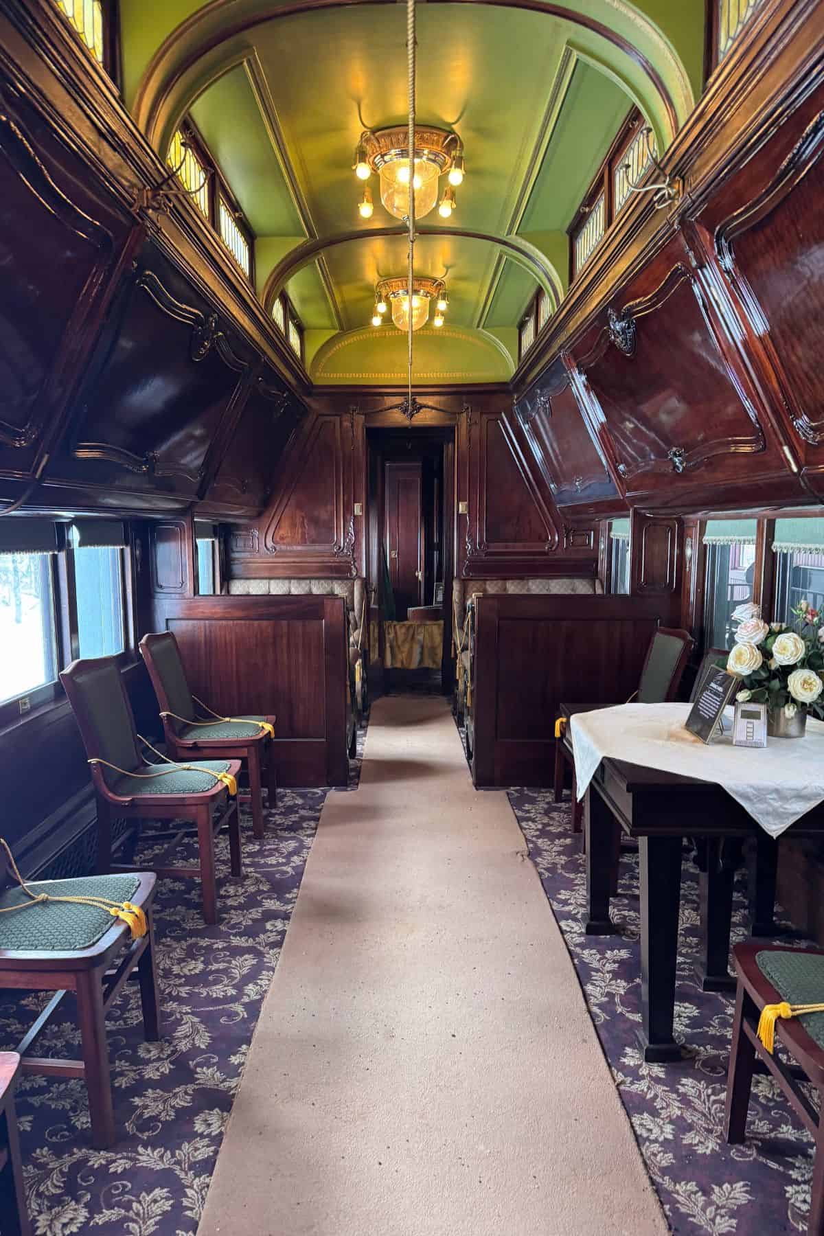 A vintage train carriage interior featuring wooden paneling, green chairs, a carpeted aisle, and a table with flowers and framed photos.