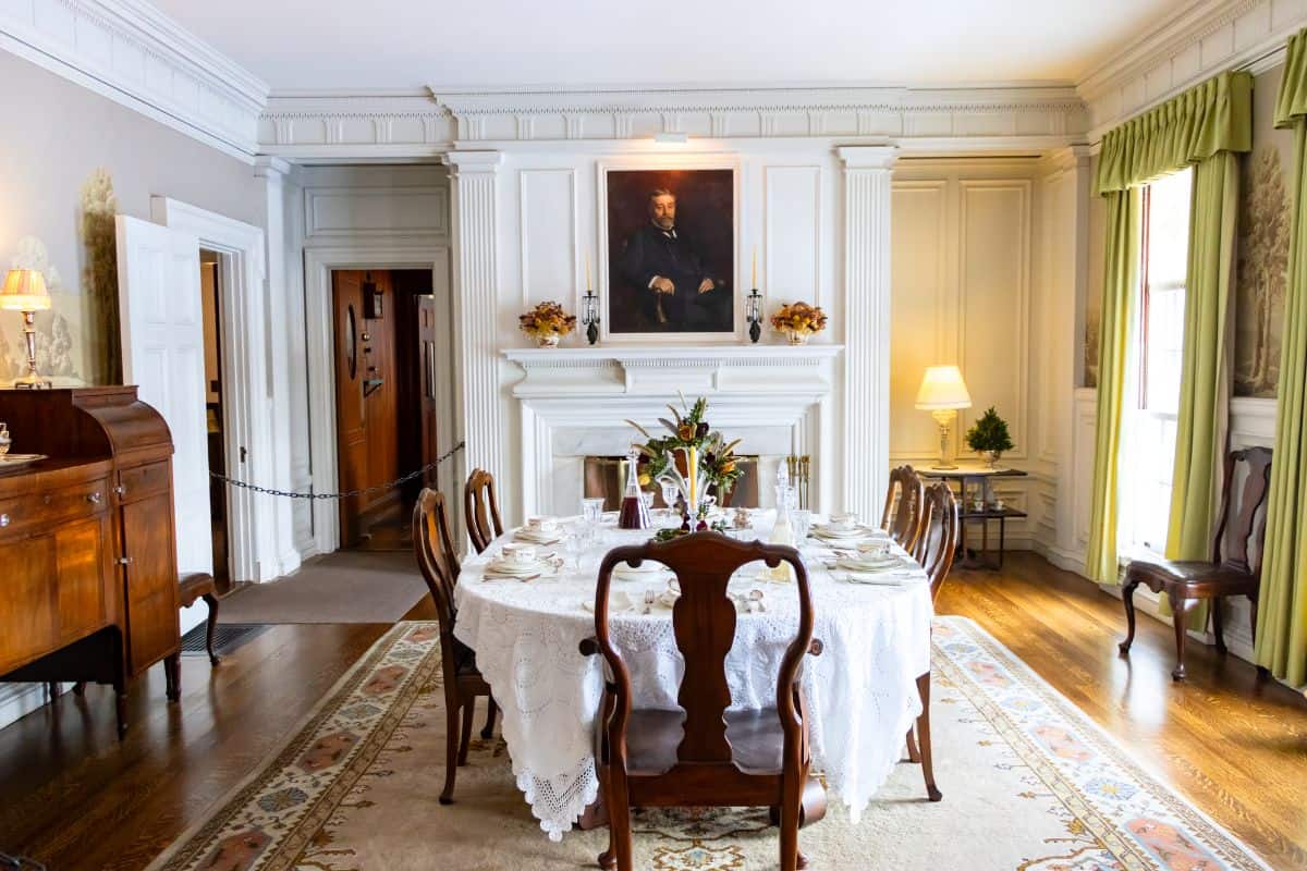 A formal dining room with a round table set for a meal, wooden chairs, a fireplace, portrait above the mantel, and large windows with green curtains.