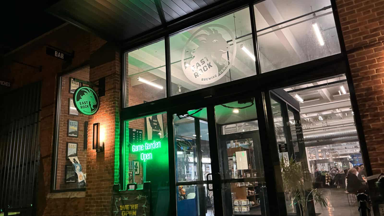 The entrance of East Rock Brewing Co. at night, with a green neon sign reading "Game Garden Open" visible through glass doors.