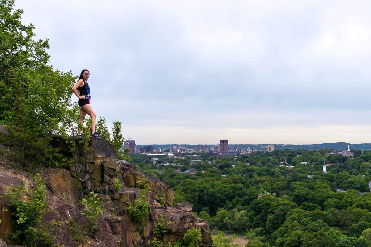 A person stands on a rocky ledge overlooking a city skyline and a forested area under a cloudy sky.
