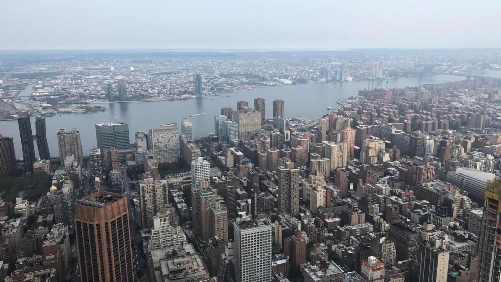 Aerial view of a densely packed cityscape with tall buildings, a wide river, and urban areas extending into the distance under a hazy sky.