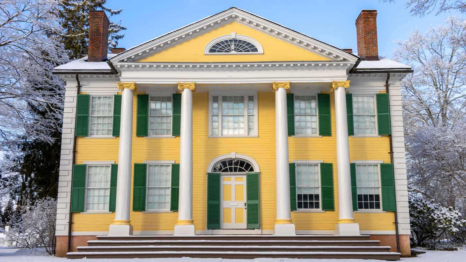 A yellow two-story house with green shutters, white columns, and a triangular pediment, surrounded by snow-covered trees and ground.