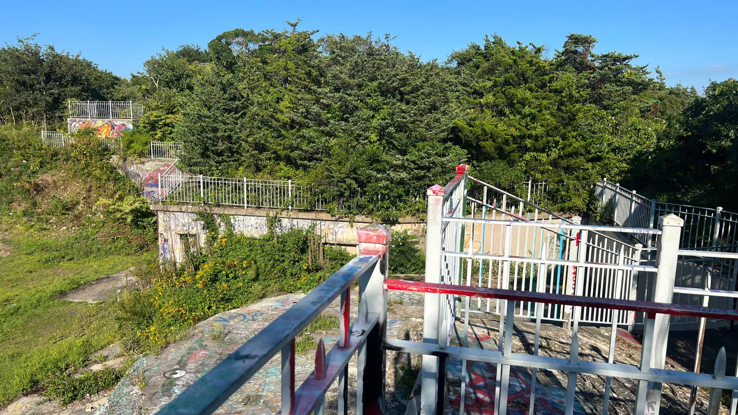 A fenced area with metal railings and gates, surrounded by trees and overgrown vegetation, with visible graffiti on the stone structures and ground.