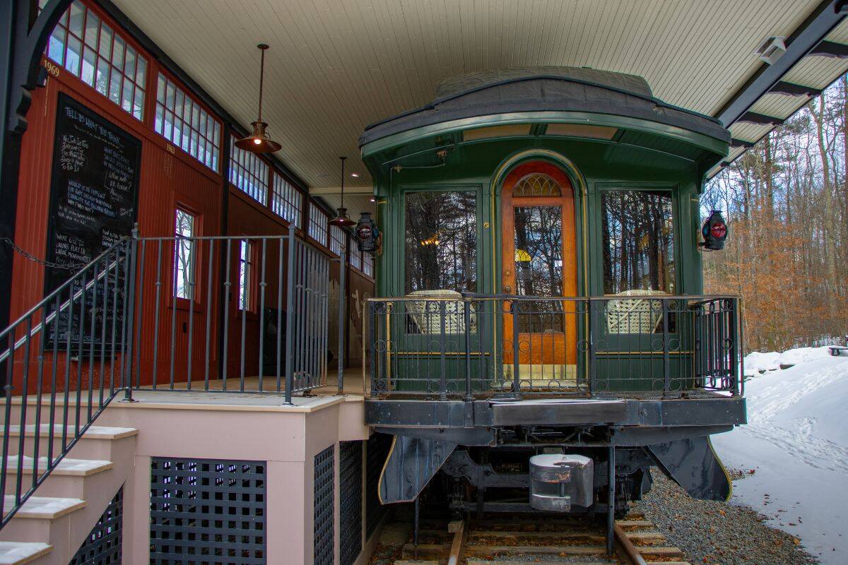 A vintage green train car is stationed at a covered platform next to a red building, with snow visible on the ground and trees in the background.