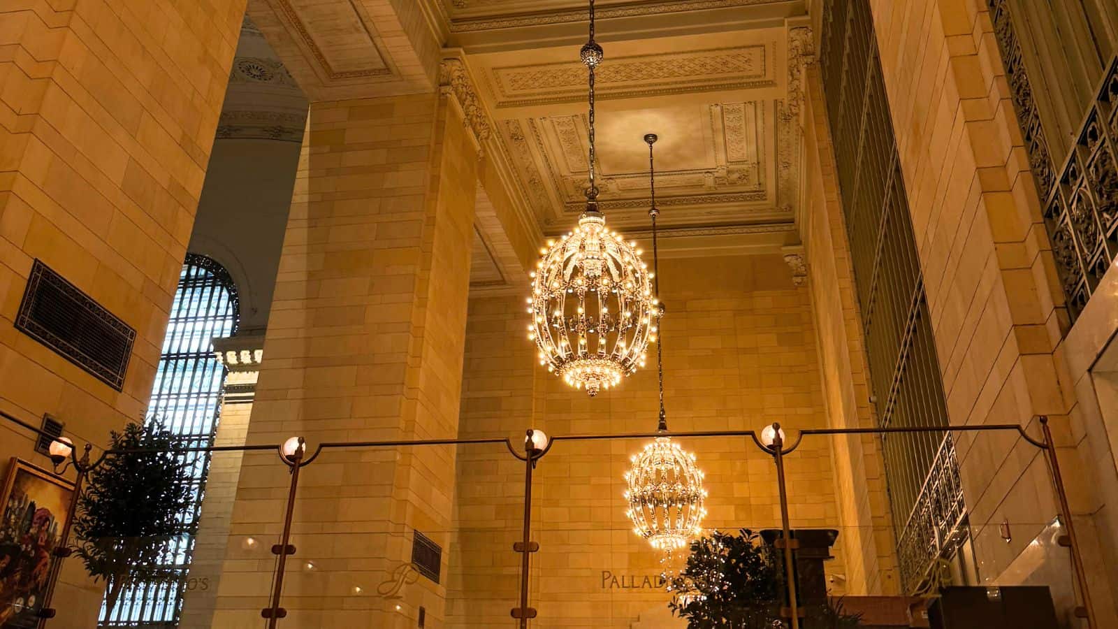 Large chandeliers hang from a high, ornate ceiling in an elegant hall with tall stone walls and arched windows.