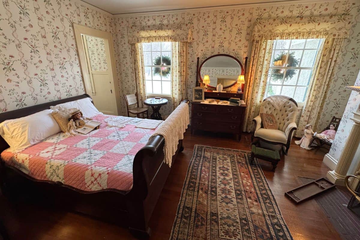 A vintage bedroom with a wooden bed, floral wallpaper, quilt, antique furniture, an armchair, and dolls near the fireplace, lit by natural light from two windows.