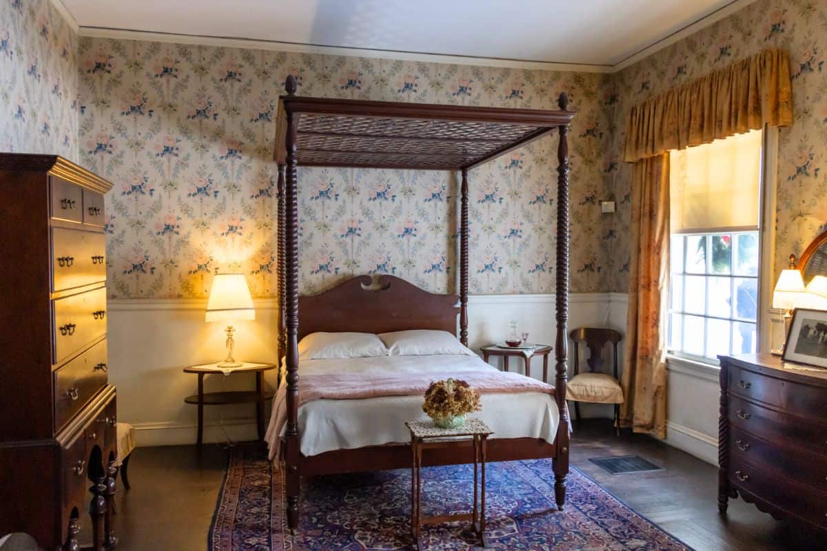 A traditional bedroom with a four-poster bed, floral wallpaper, wooden furniture, two lamps, and a window with gold curtains.