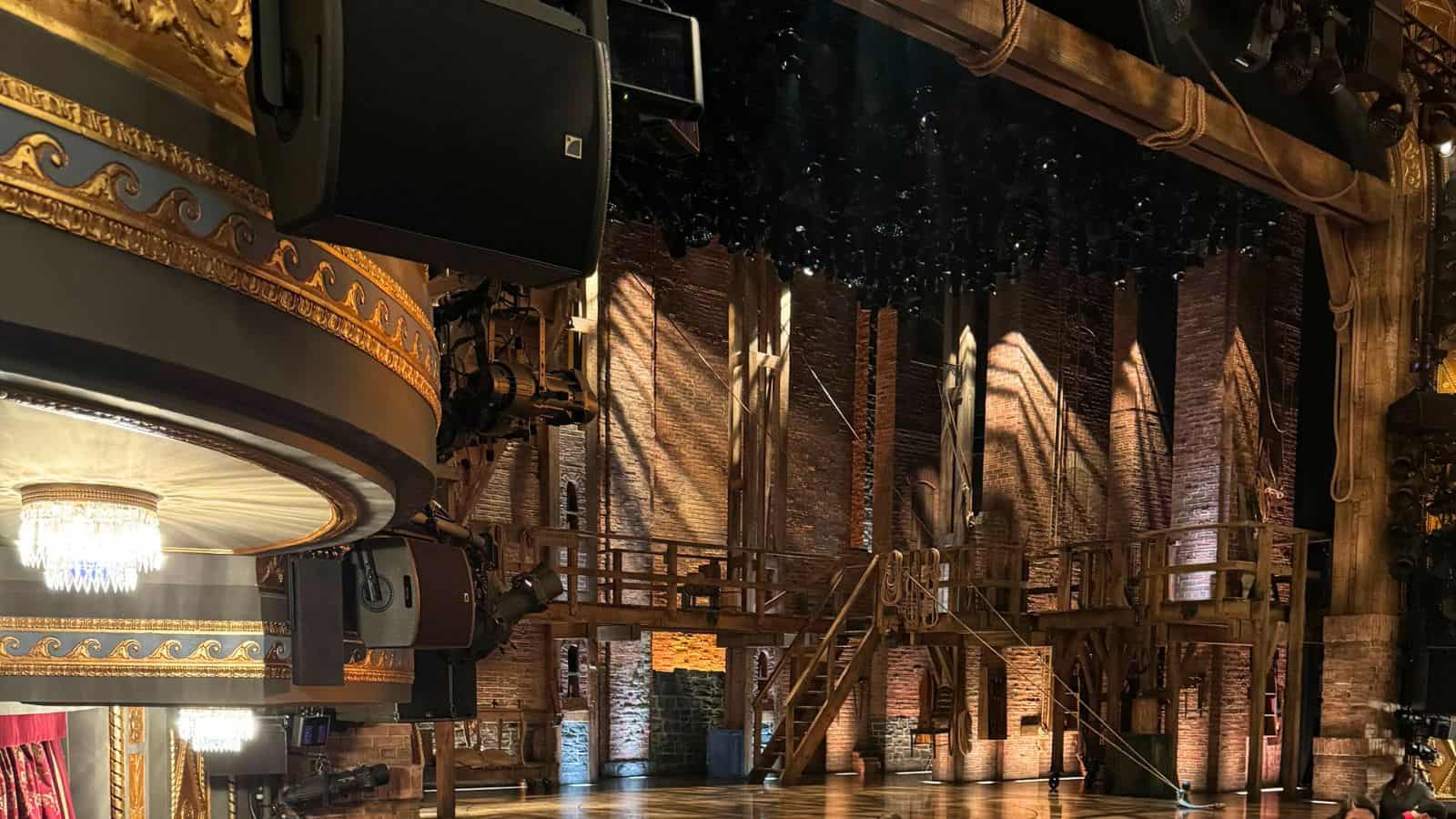 Empty stage set with wooden scaffolding, exposed brick walls, and theatrical lighting in a historic theater with ornate gold detailing.