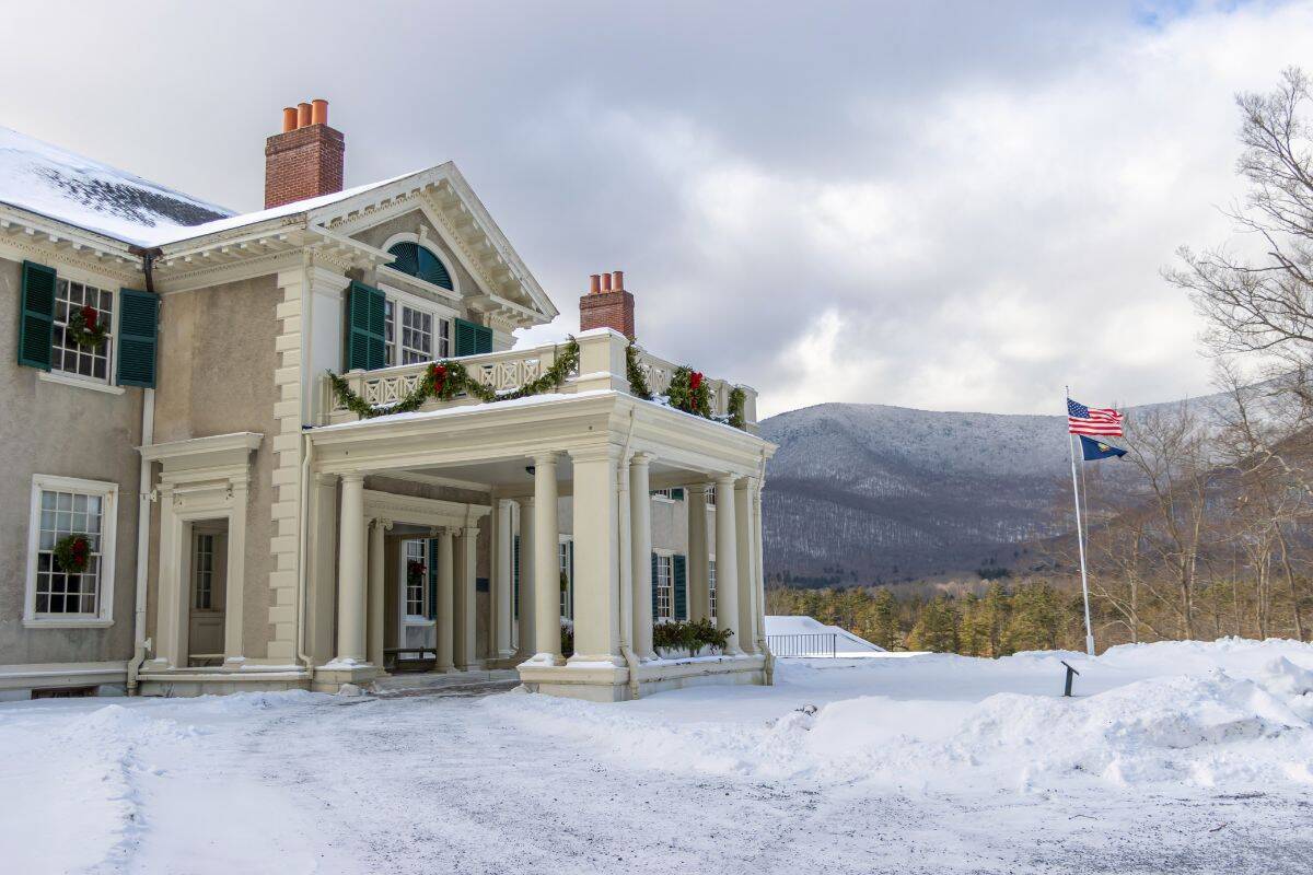 A large historic house with columns and green shutters is surrounded by snow, with mountains in the background and two flags flying on a flagpole.