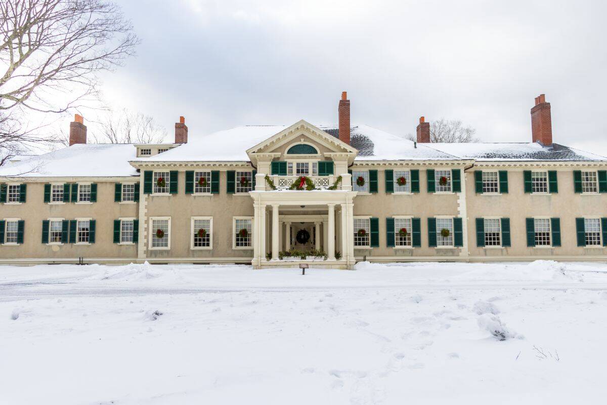 Large historic mansion with green shutters, decorated with wreaths, and surrounded by snow under an overcast winter sky.