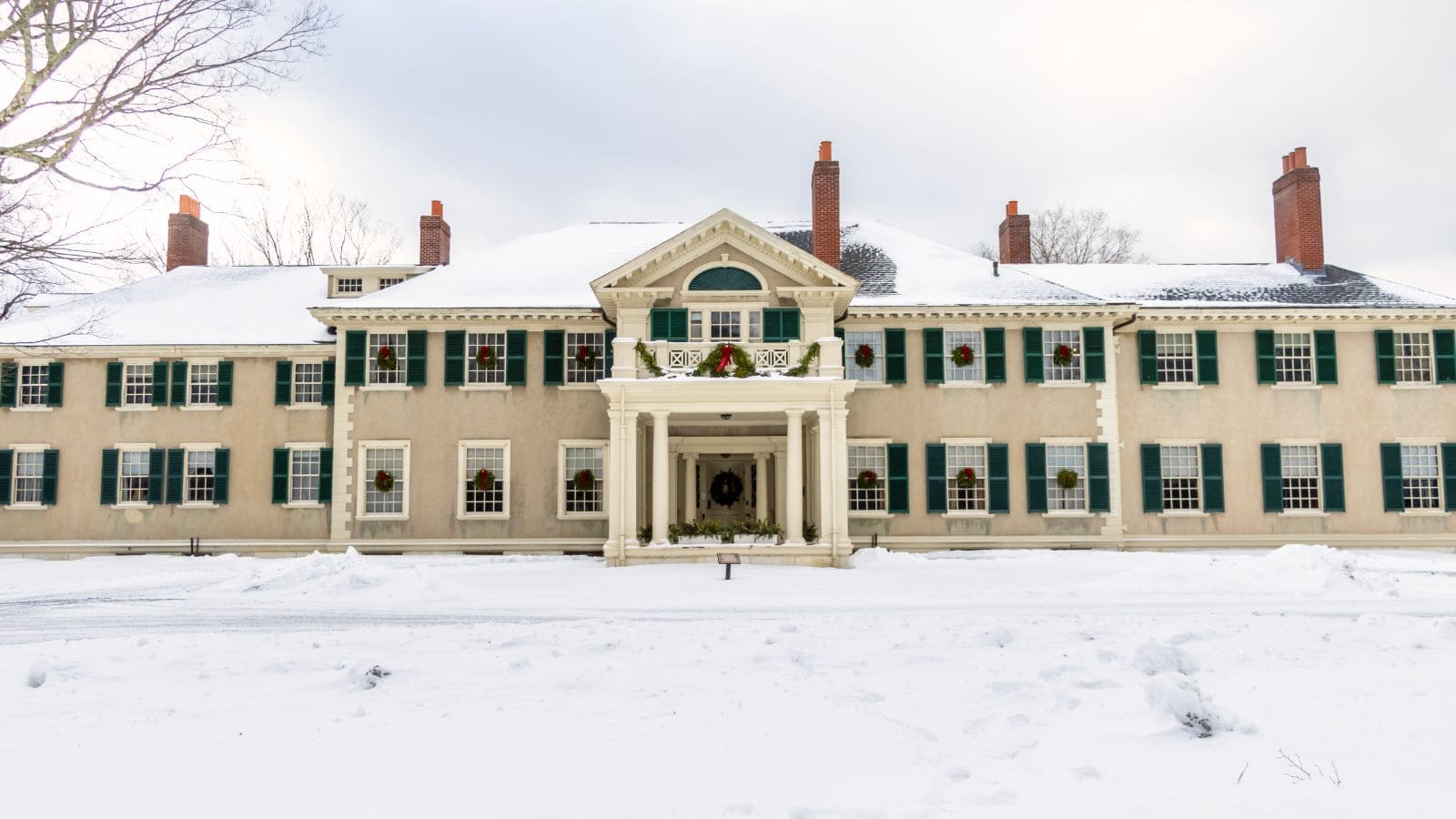 Large colonial-style house with green shutters, brick chimneys, and snow covering the roof and ground on a cloudy winter day.