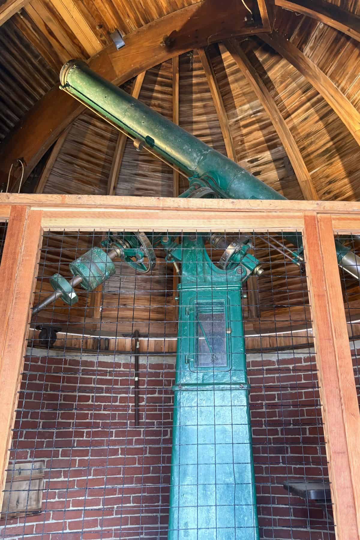 A large green telescope is mounted inside a small brick observatory with a wooden domed roof, behind a wire and wood barrier.
