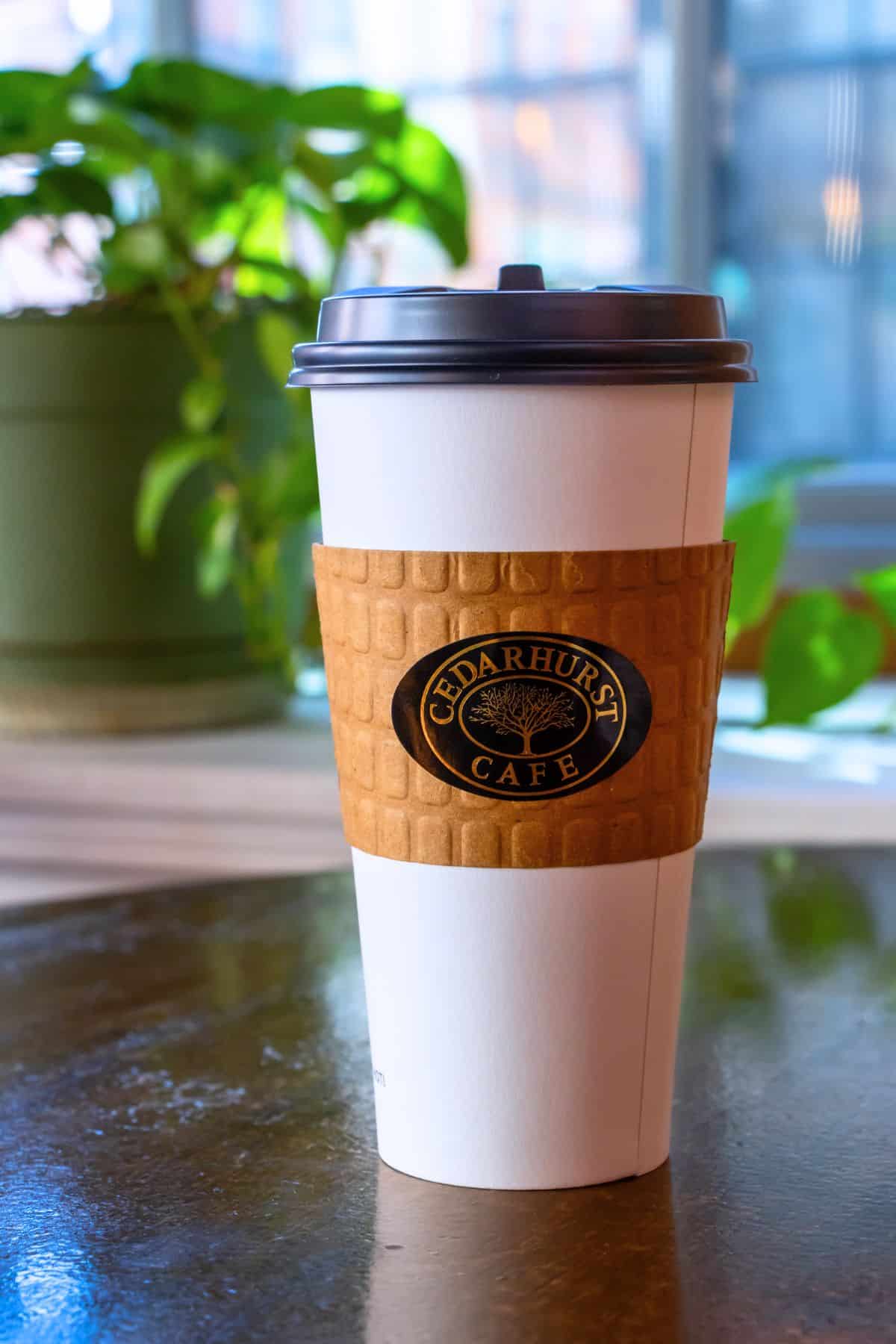 A takeaway coffee cup with a Cedarhurst Cafe sleeve sits on a table, with potted plants and a window in the blurred background.