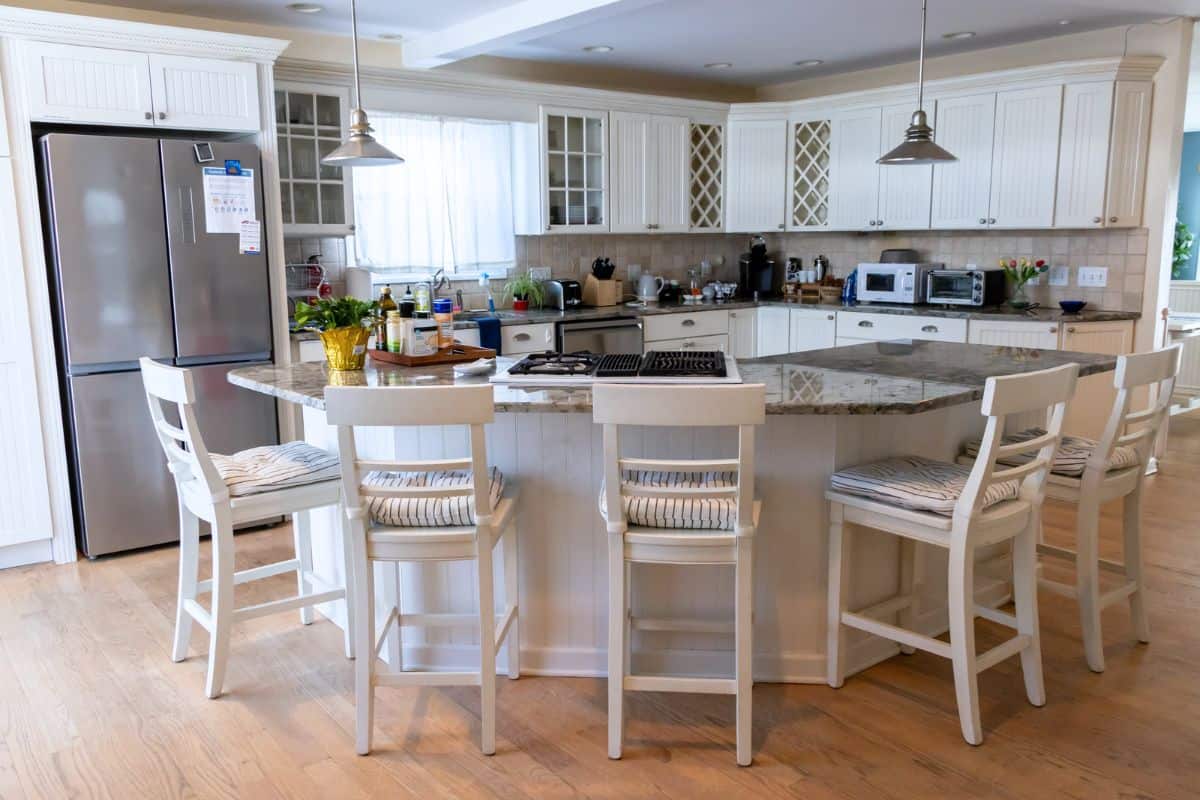 Modern kitchen with white cabinets, granite island, four white chairs, stainless steel appliances, and various kitchen items on the counters.