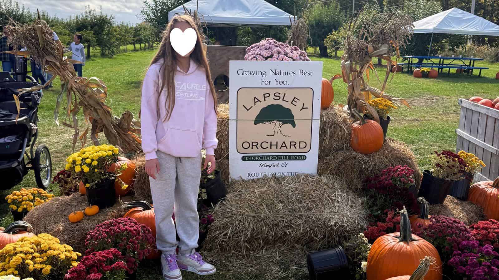 A person stands in front of a Lapsley Orchard sign surrounded by hay bales, pumpkins, and flowers at an outdoor autumn event.