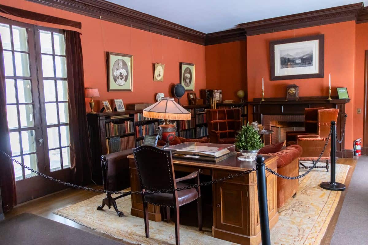 A vintage study room with an ornate desk, leather chair, bookshelves, framed portraits, and a fireplace, separated by a rope barrier.