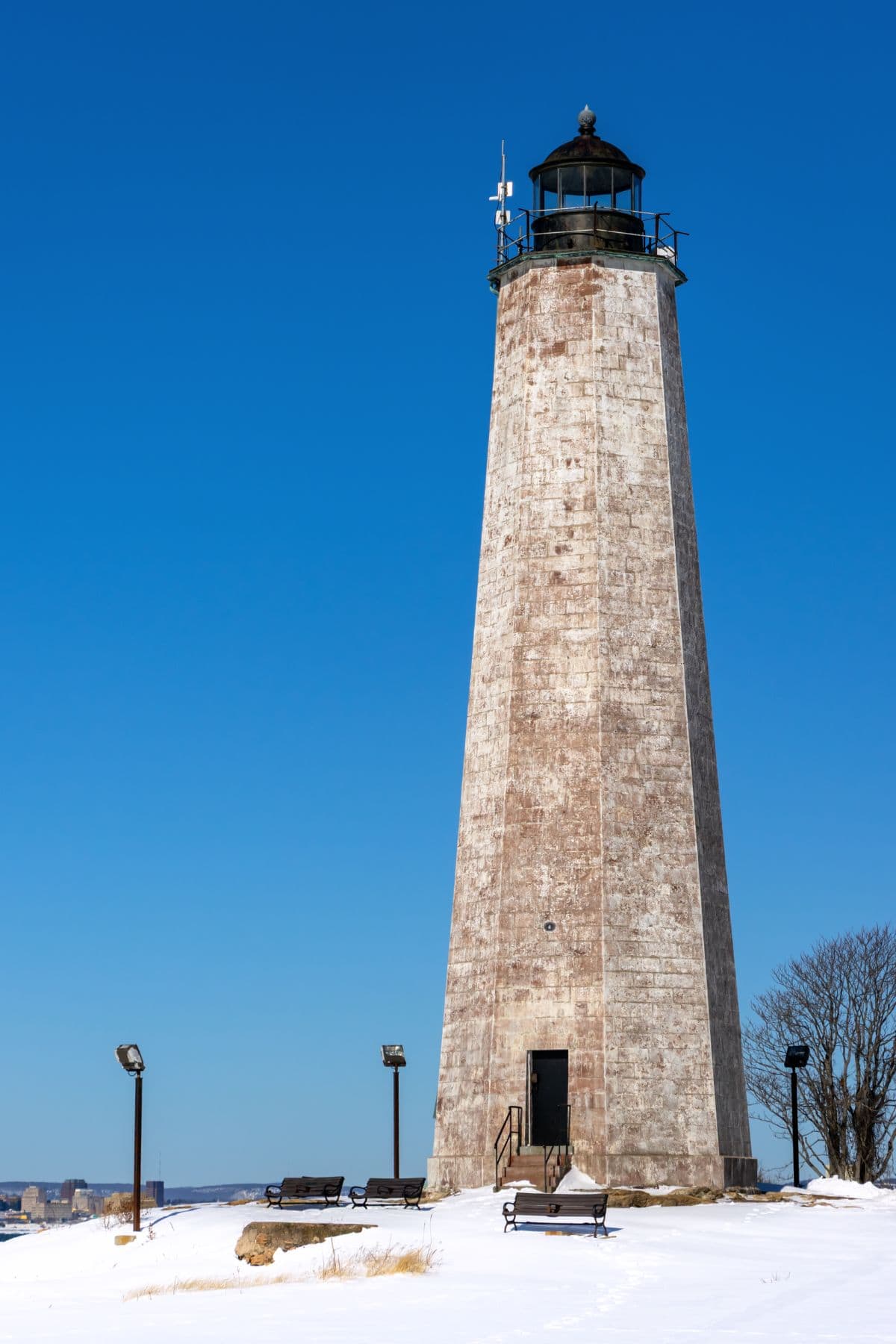 A tall, weathered lighthouse stands on a snowy landscape under a clear blue sky, surrounded by benches and lamp posts.