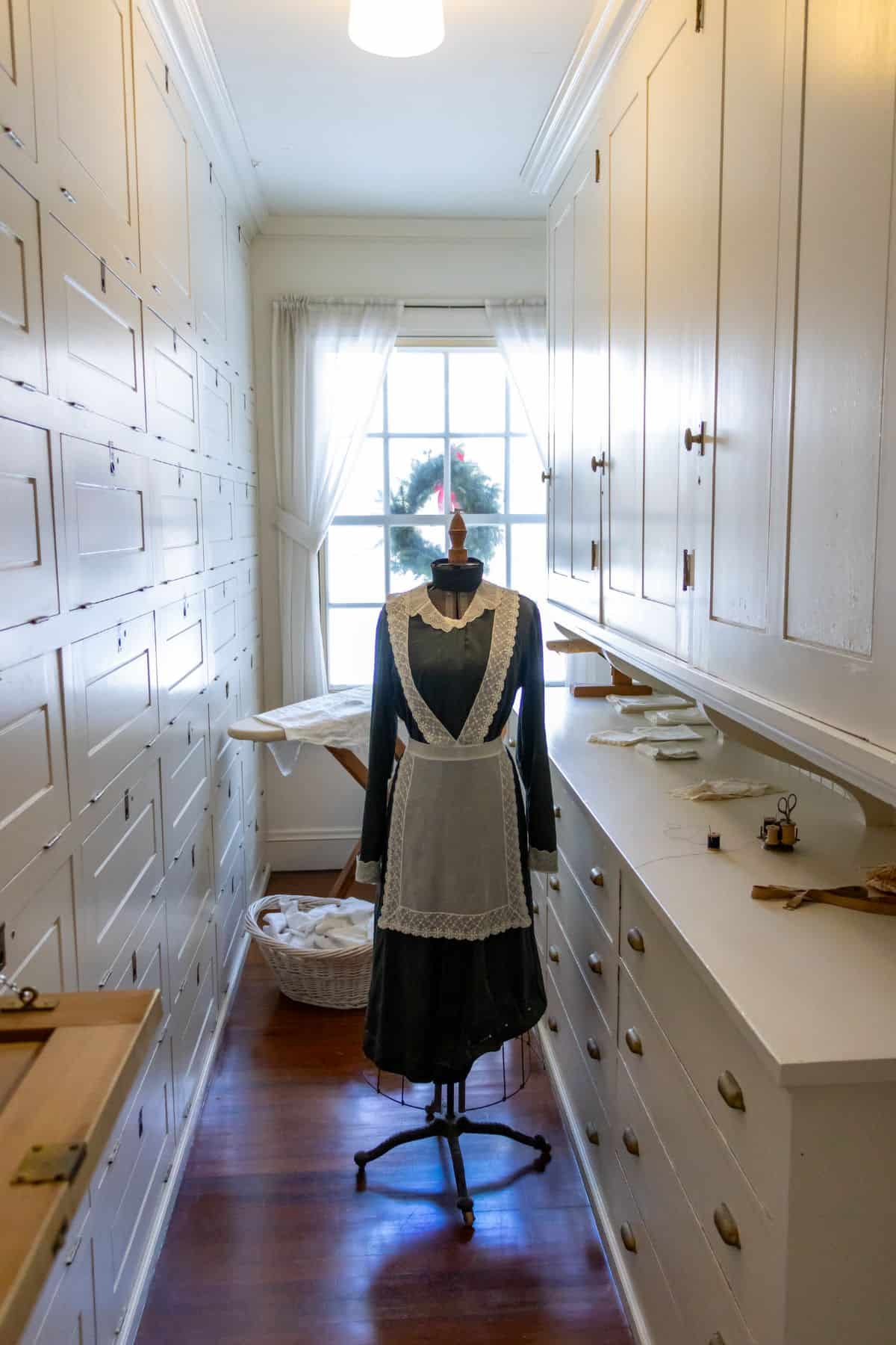 A mannequin dressed in a black dress with a white apron stands in a narrow room filled with white cabinets and drawers, with sunlight coming through a window at the end.