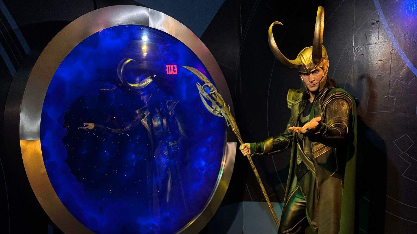 A person in a Loki costume stands beside a circular portal with their reflection visible inside, holding a staff and gesturing with one hand.