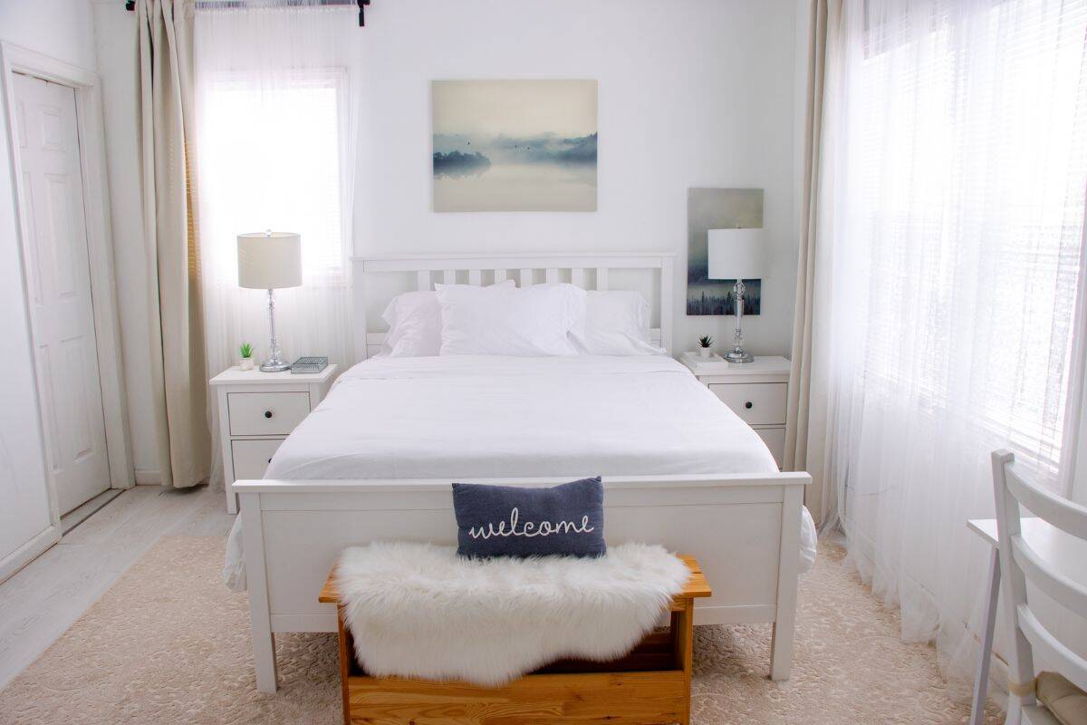 A bright, white bedroom with a neatly made bed, two nightstands with lamps, wall art above the bed, and a bench with a “welcome” pillow at the foot.