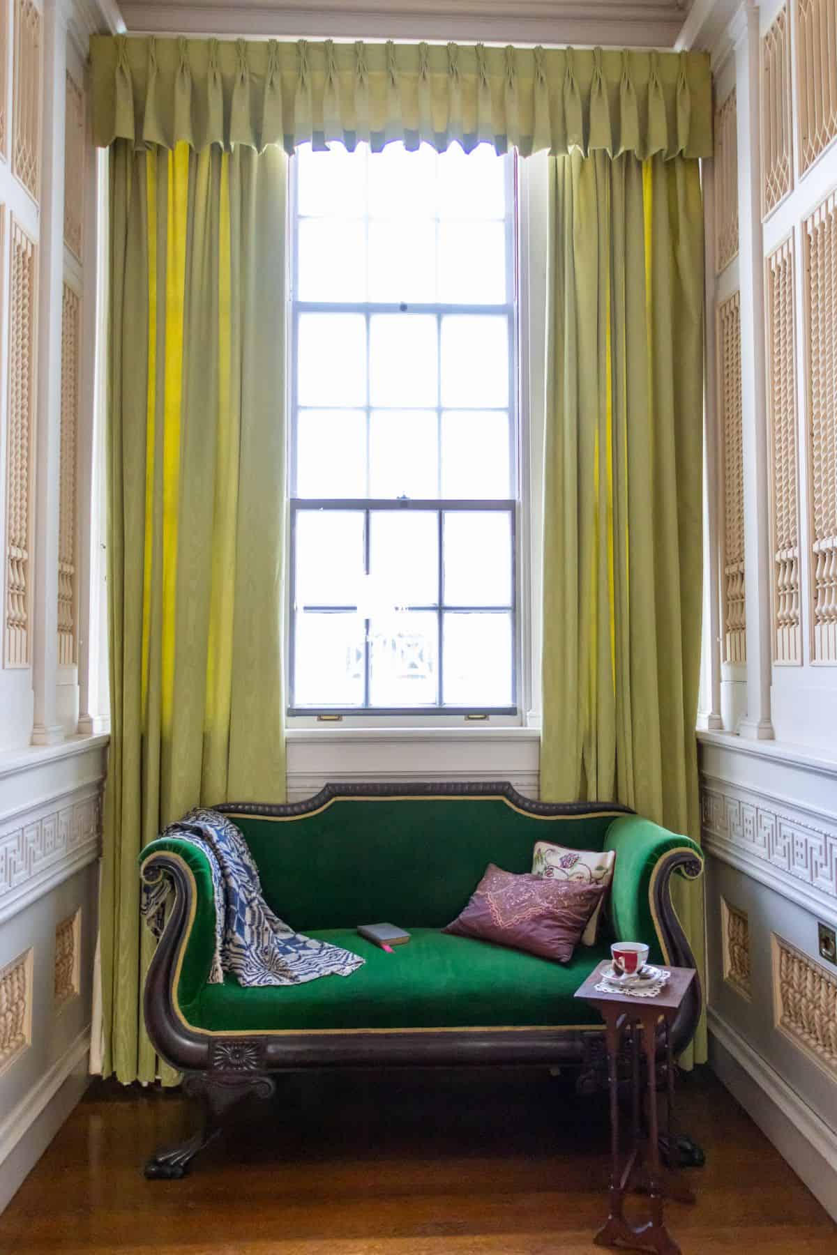 A green velvet sofa sits in front of a large window with yellow-green curtains; a small wooden side table holds a cup and saucer.
