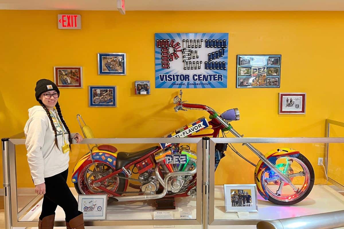 A woman in a winter hat and scarf stands beside a colorful custom motorcycle displayed in a visitor center with framed photos and signage on a yellow wall.