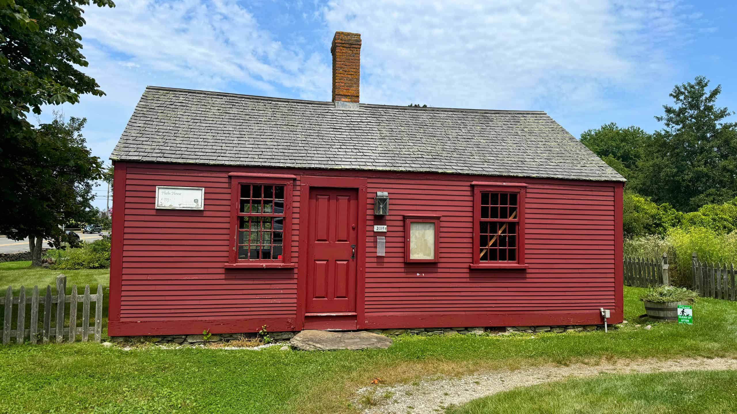 A small, red wooden house with a shingled roof and central brick chimney sits in a grassy yard with a picket fence and a clear, partly cloudy sky overhead.
