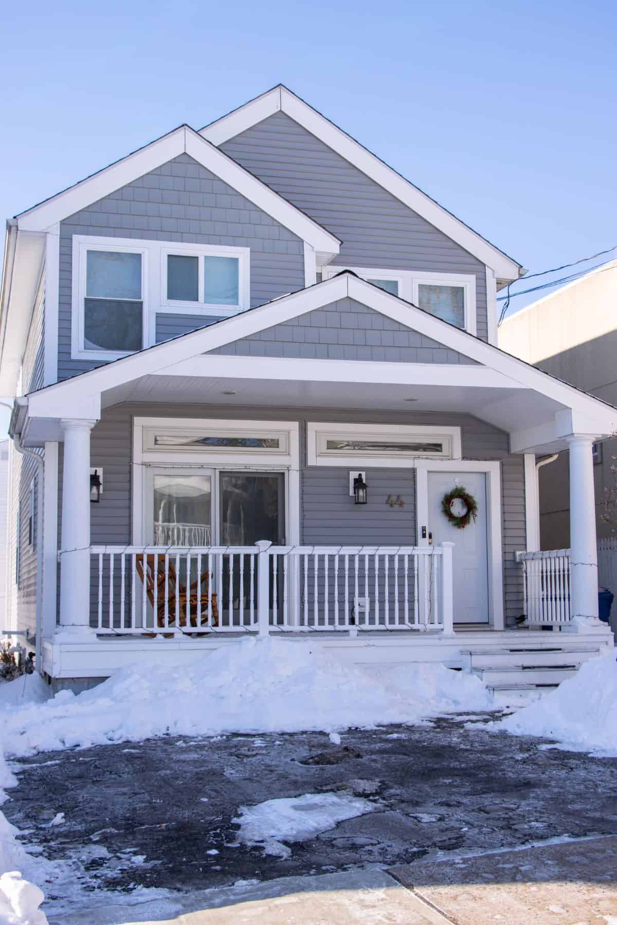 A two-story gray house with white trim, a front porch, and snow covering the ground and driveway.