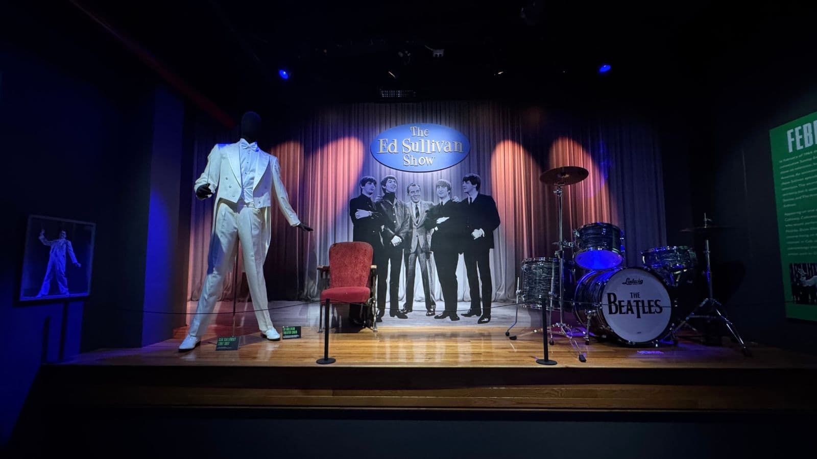 A display featuring a white suit on a mannequin, a drum set labeled "The Beatles," and a backdrop of The Beatles’ Ed Sullivan Show performance.