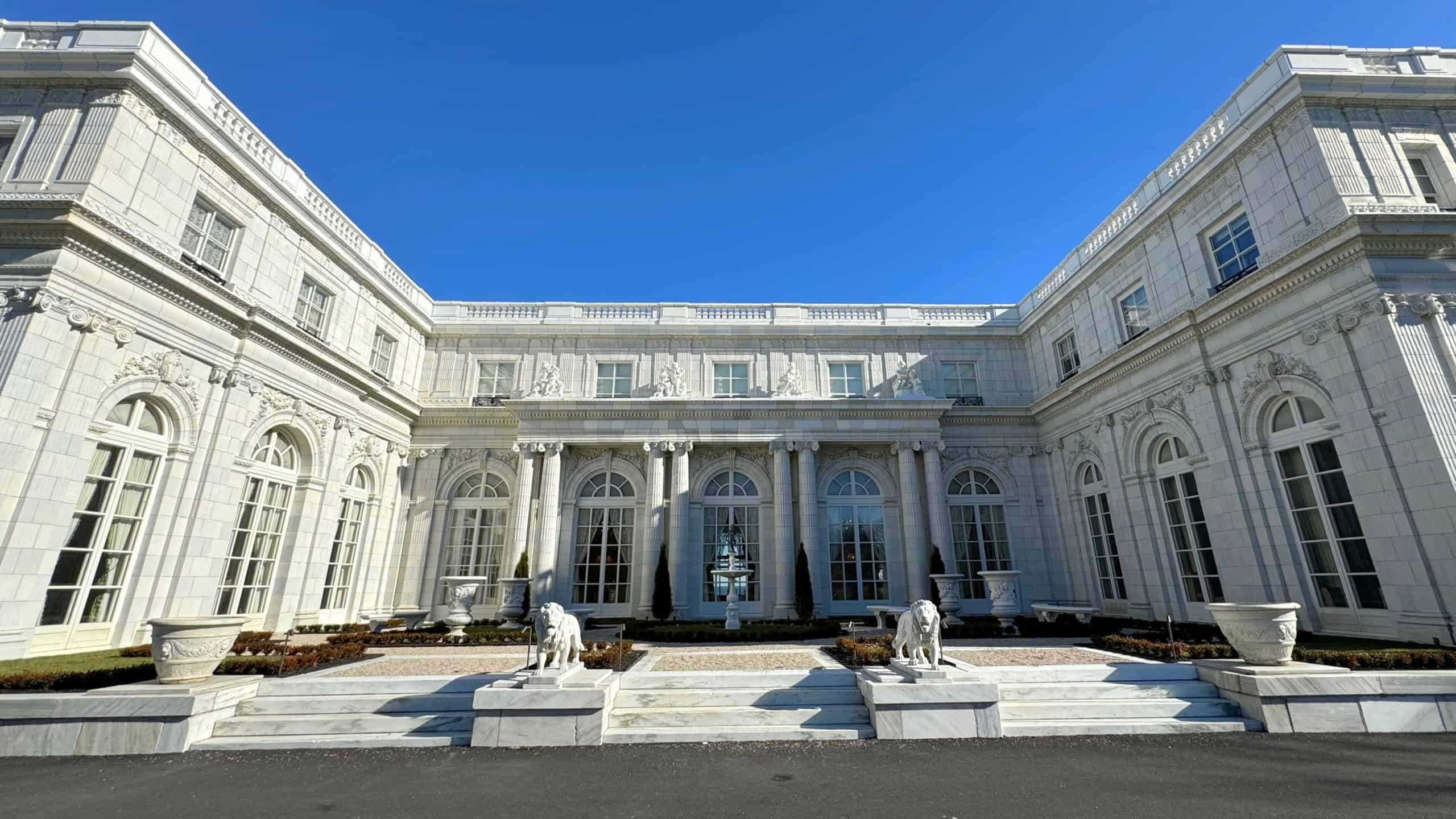A large white neoclassical building with arched windows, columns, and two stone lion statues flanking the entrance under a clear blue sky.